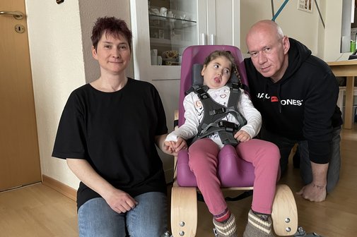 Familie Heubner Das Ehepaar Heubner kniet links und rechts neben ihrer Tochter Nena am Boden, das Mädchen sitzt in dem speziellen Therapiestuhl, den der VdK durchsetzen konnte