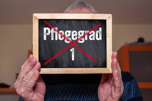 Pflegegrad 1 Fotomontage: Ein älterer Mann hält ein Schild mit dem durchgestrichenen Schriftzug: Pflegegrad 1. Das Symbolbild steht für die Abschaffung des Pflegegrads 1.