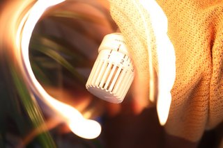Das Bild zeigt ein Heizungsthermostat und eine Wolldecke die über der Heizung liegt. Ein Kranz aus hellem Licht durchzieht das Bild.