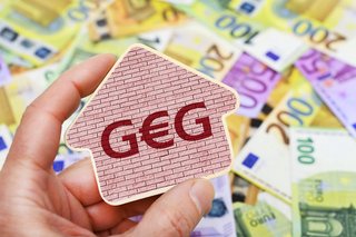 Eine Hand hält ein Schild mit der Aufschrift "GEG", da E ist ein Eurozeichen. Dahinter liegen viele Euro-Scheine. |