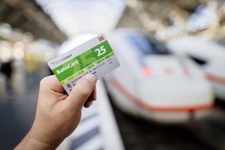Eine Person steht an einem Bahngleis, an dem ein ICE der Deutschen Bahn steht, sie hält eine Bahncard 25 in der Hand. 