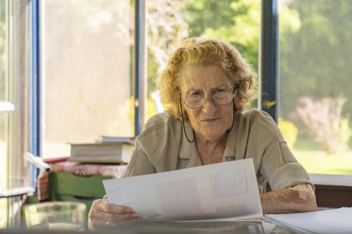 Abrechnung des Pflegedienstes Konzentrierte ältere Frau mit Brille, die an ihrem Schreibtisch in einem sonnendurchfluteten Raum mit Blick ins Grüne sitzt und Papierkram durchgeht, möglicherweise Rechnungen oder medizinische Unterlagen