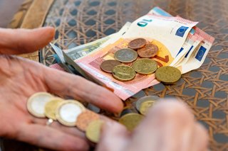 Hand einer älteren Frau mit Euromünzen, auf dem Tisch davor liegen Euroscheine und -münzen 