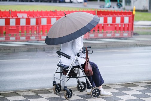 Rente und Alter Eine Frau mit aufgespanntem Regenschirm sitzt auf ihrem Rollator auf einem Platz. Im Hintergrund ist ein Baustellenzaun zu sehen.