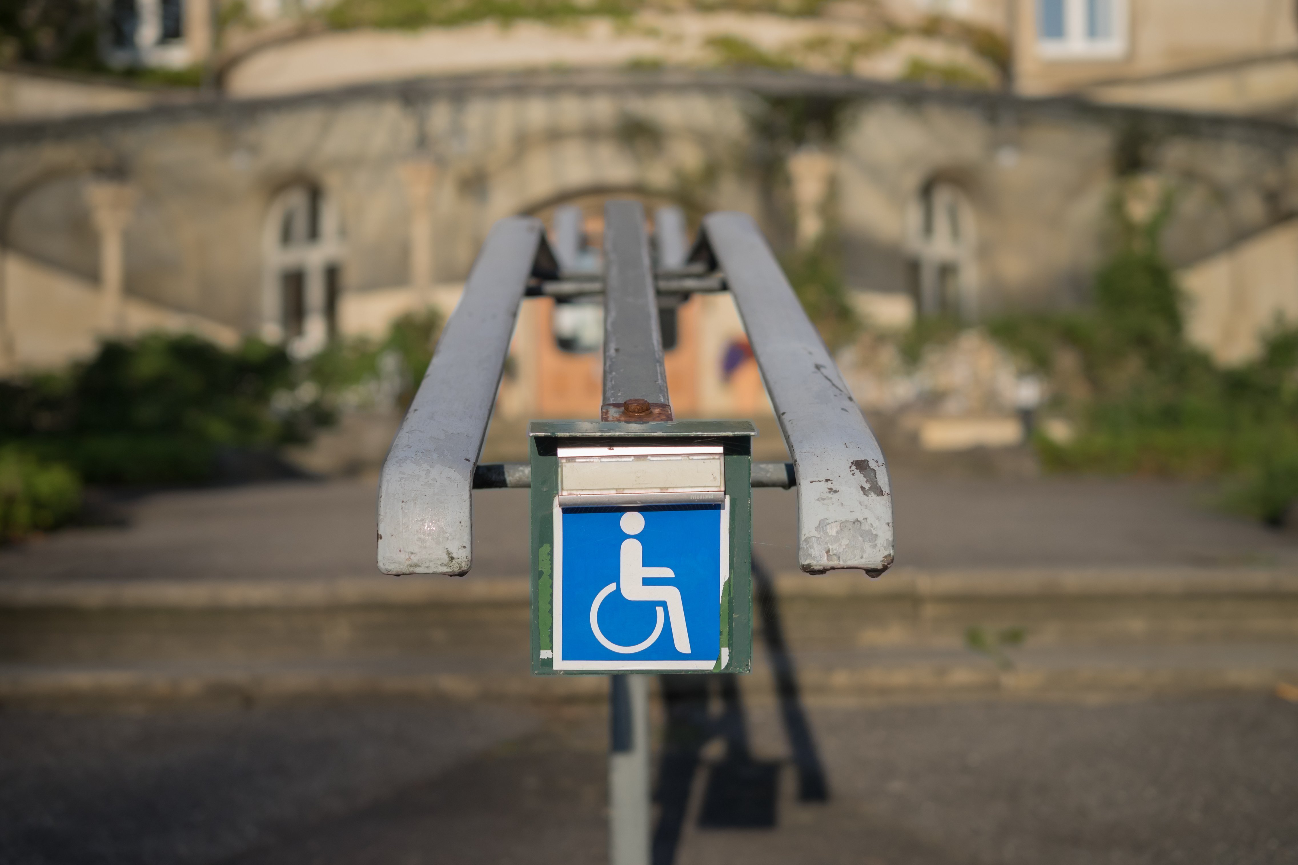 Barrieren in der Stadt Eine Stufe mit einem Handlauf. An diesem ist eine Klingel mit einem Rollstuhl-Symbol angebracht; Symbol für eine nicht barrierefreie Stelle im öffentlichen Raum.