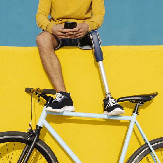 Leben mit Behinderung Ein Mann mit einer Beinprothese sitzt auf einer Mauer und schaut auf sein Smartphone. Seine Füße hat er auf ein Fahrrad gestützt, das an der Mauer lehnt.