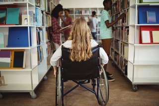 Junge Frau sitzt in einem Rollstuhl. Sie befindet sich in einer Bibliothek.