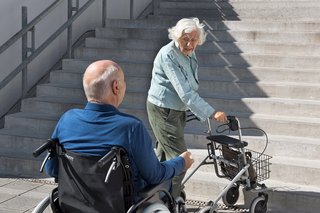 Ein älterer Mann im Rollstuhl und eine ältere Frau mit Rollator stehen am Fuß einer unüberwindbaren Treppe.
