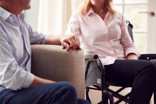 Älterer Mann auf einem Sofa und ältere Frau im Rollstuhl sitzen vertraut nebeneinander, halten sich an der Hand. 