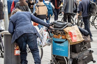 Flaschensammlerin in der Innenstadt von München - eine Frau mit einem Einkaufswagen voller gesammelter Flaschen und anderer Dinge beugt sich über einen Mülleimer, schaut hinein.