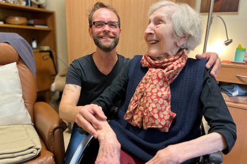 Besuch im Seniorenheim Dirk Angele und Anita Göbels, die Bewohnerin des Seniorenheims, in dem Dirk Angele ehrenamtlich tätig ist. Sie sitzt im Rollstuhl, er sitzt hinter ihr, legt eine Hand auf ihre Schulter und die andere auf ihren Arm.