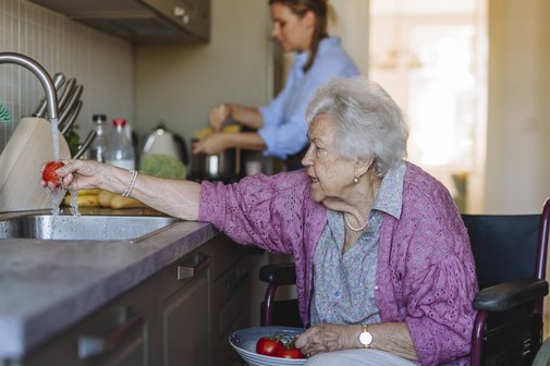 Angehörigenpflege Ältere Frau im Rollstuhl in der Küche wäscht Obst unter dem Wasserstrahl der Spüle. Im Hintergrund sieht man eine jüngere Frau, die in der Küche etwas erledigt.