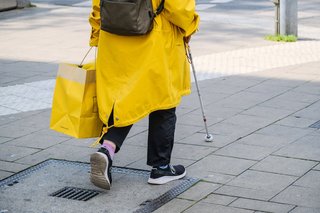 Person in gelben Regenmantel läuft mit einem Blindenstock entlang eines Blindenleitsystems 
