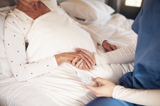 Hand einer Pflegerin hält die einer älteren, bettlägrigen Patientin