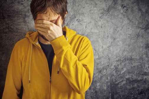 Psychische Erkrankung Erwerbsminderung Ein Mann in gelber Sweatjacke steht vor einer grauen Wand, er hält sich eine Hand vor die Augen, senkt den Kopf