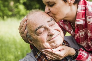 Ein Frau küsst einen älteren Mann liebevoll auf die Stirn, hält sein Gesicht in den Händen.