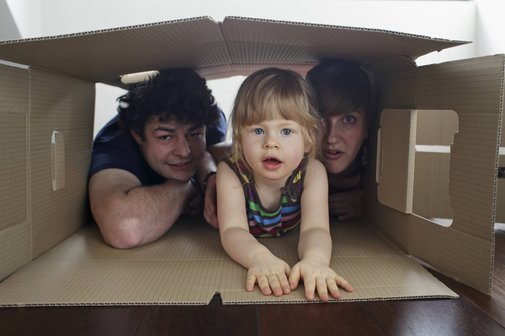 Wohnkostenlücke im Bürgergeld Eine junge Familie: Vater, Mutter und ein kleines Mädchen schauen gemeinsam aus einem großen Pappkarton heraus, der auf dem Boden liegt.