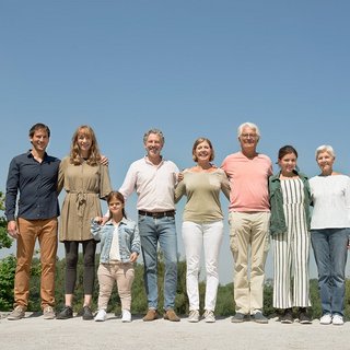 Menschen verschiedenen Alters mit und ohne Behinderung stehen untergehakt und lächelnd in einer Reihe in der Sonne