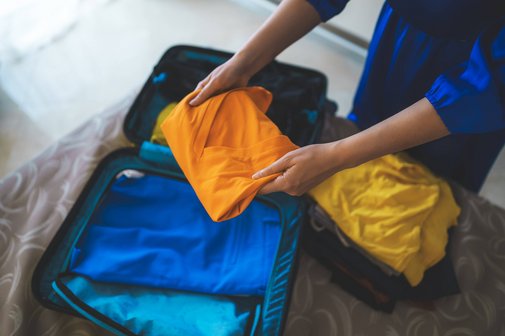 Koffer packen Eine Frau packt Kleidungsstücke in einen kleinen Koffer, der auf einem Bett liegt.