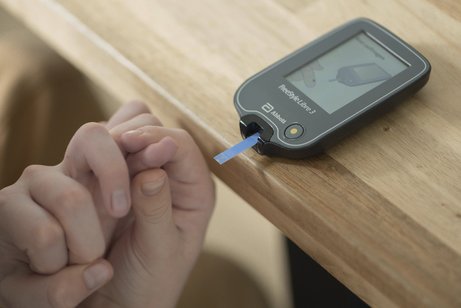 Blutzuckermessung bei Kind mit Diabetes Ein Blutzucker-Messgerät mit Teststreifen liegt auf einem Holztisch, eine Kinderhand hält einen Finger fest und führt ihn zum Messgerät.