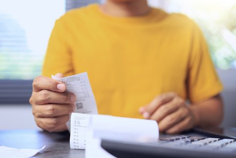 Steigende Kosten Eine Person im gelben Pullover sitzt an einem Schreibtisch, vor sich einen Taschenrechner und ein Dokument, wahrscheinlich eine Rechnung, als Symbol für steigende Kosten