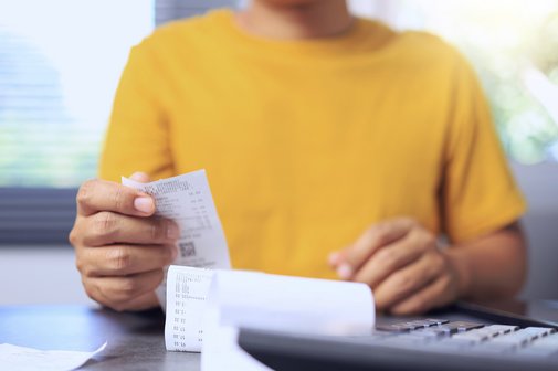 Steigende Kosten Eine Person im gelben Pullover sitzt an einem Schreibtisch, vor sich einen Taschenrechner und ein Dokument, wahrscheinlich eine Rechnung, als Symbol für steigende Kosten