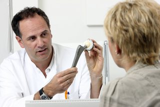 Szene in einer Arztpraxis zeigt einen Arzt im Gespräch mit einer Patientin. Erklärt ihr die Funktionsweise eines künstlichen Hüftgelenkes vor einer Operation