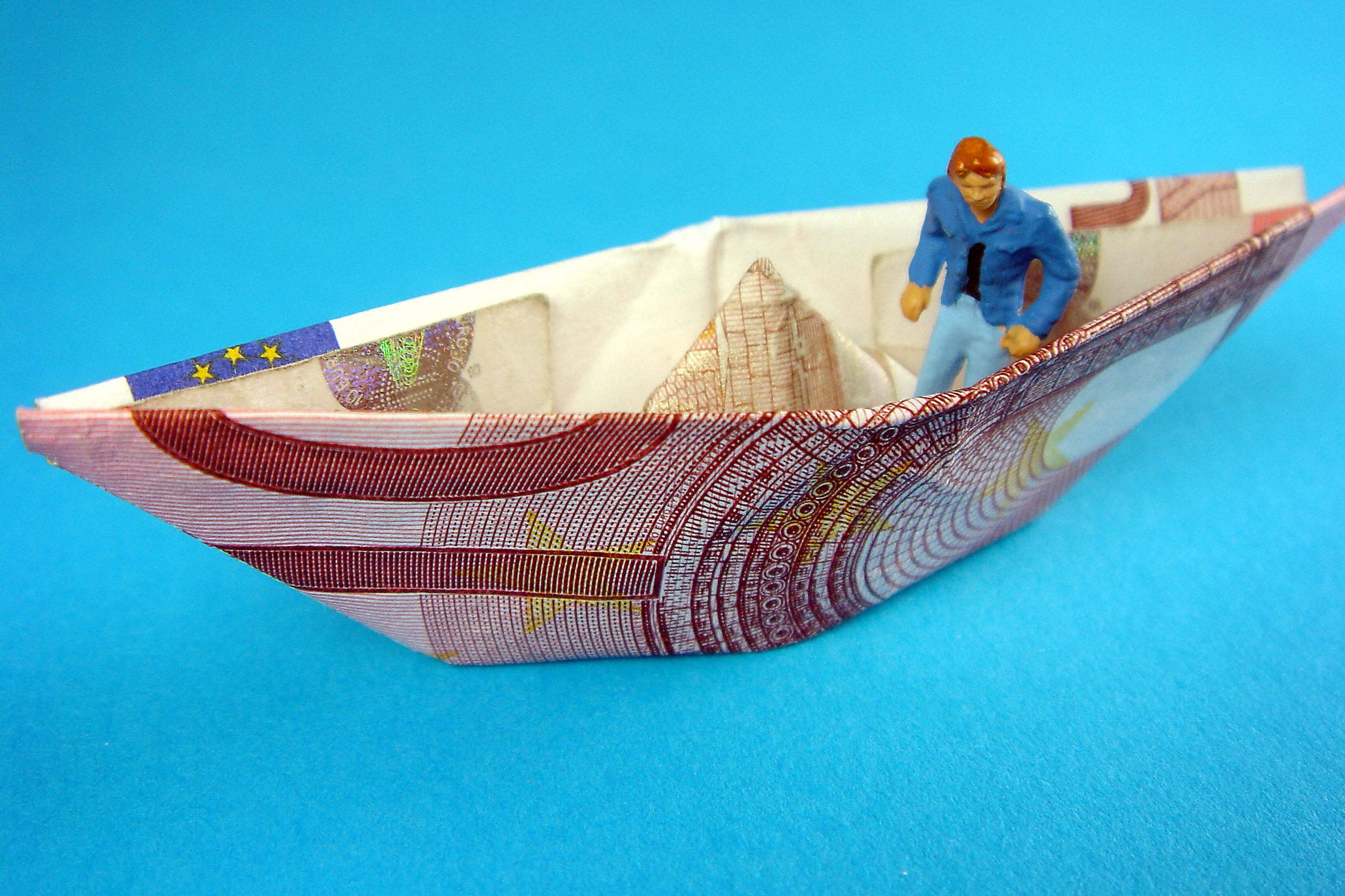 Das Symbolbild zeigt ein Papierschiffchen, das aus einem Geldschein gefaltet ist. Darin sitzt ein kleines Figürchen.