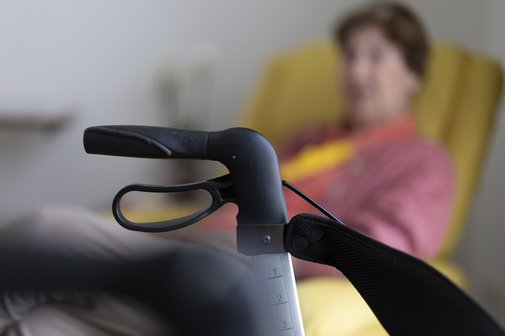 Frau mit Rollator Seniorin sitzt in ihrem Zimmer im Seniorenheim hinter einem Rollator in einem gelben Sessel. Der Rollator im Bildvordergrund ist scharf zu sehen, die Seniorin im Hintergrund verschwommen.