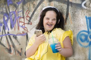 Junge Frau mit Downsyndrom, sie lacht in die Kamera, hält ihr Smartphone in der einen Hand und einen Plastikbecher mit einem Getränk in der anderen. Sie trägt Kopfhörer.