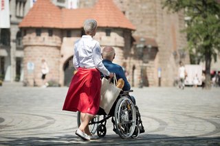 Eine Seniorin schiebt ihren Mann, der im Rollstuhl sitzt, über einen Platz, man sieht im Hintergrund ein altes Stadttor.