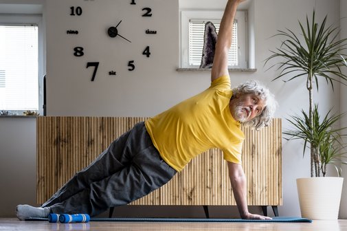 Bewegung und Fitness als Prävention Ein älterer Mann im gelben T-Shirt macht eine Fitnessübung auf einer Sportmatte, er stemmt sich zu einer seitlichen Planke hoch.