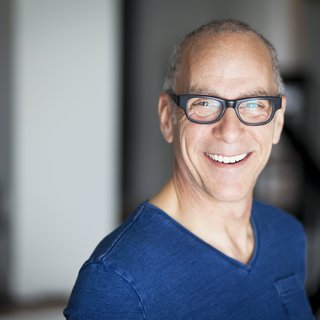 Mann mittlerem Alters mit Brille und blauem Pullover lacht in die Kamera