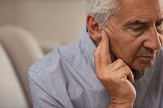 _Seitenansicht eines älteren Mannes mit Symptom eines Hörverlusts sitzt auf der Couch mit den Fingern in der Nähe des Ohrs
