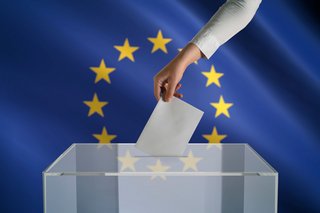 Au dem Foto sieht man eine Wahlurne, in die jemand einen Wahlzettel einwirft. Im Hintergrund ist die Europaflagge zu sehen.
