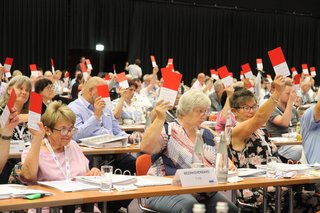 Delegierte an langen Tischen halten rote Stimmkarten hoch. 