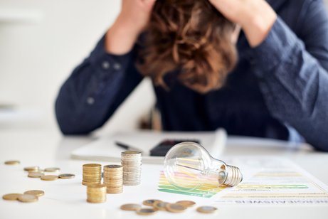 Energiearmut Symbolfoto für Energieschulden: Ein junger Mann sitzt an einem Tisch, auf dem Münzen, Schreiben und eine Glühbirne liegen. Er hat den Kopf gesenkt, rauft sich die Haare.