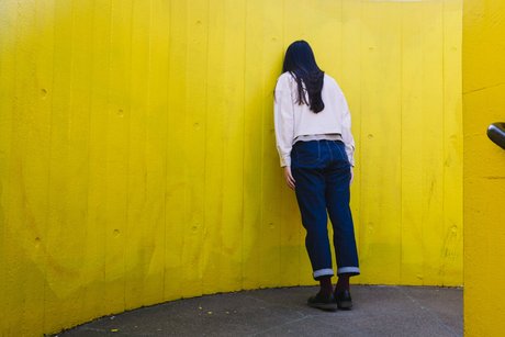 Eine junge Frau lehnt an einer gelben Wand mit dem Gesicht zur Wand, ihre Körperhaltung wirkt niedergeschlagen. 