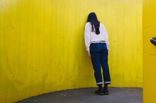 Eine junge Frau lehnt an einer gelben Wand mit dem Gesicht zur Wand, ihre Körperhaltung wirkt niedergeschlagen. 