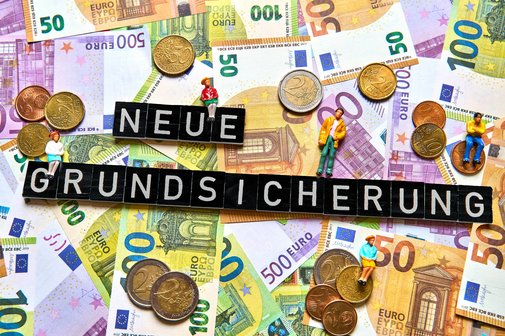 Neue Grundsicherung Geldscheine und Münzen, darauf bilden Buchstabenwürfel die Worte "Neue Grundsicherung"