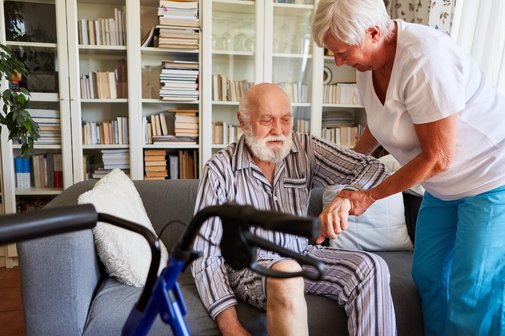 Pflege durch Angehörige Ein älterer Mann im gestreiften Pyjama sitzt auf einem Sofa. Eine ältere Frau hält ihn am Arm und hilft ihm aufzustehen. Im Vordergrund steht ein Rollator.