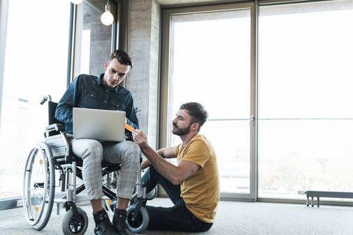 Gleichstellung mit schwerbehinderten Menschen Ein junger Mann im Rollstuhl im Büro, neben ihm sitzt ein Arbeitskollege auf dem Boden, sie sprechen und schauen gemeinsam auf ein Notebook