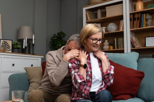 Anstrengende Angehörigenpflege Ein älterer Mann umarmt eine ältere Frau von hinten, sie sitzen auf einem Sofa. Er hat sein Gesicht von hinten in ihrer Schulter vergraben. Sie hält seine Hände, blickt geradeaus mit einem angestrengten, ernsten Ausdruck. Symbolfoto für Erschöpfung in der Angehörigenpflege.
