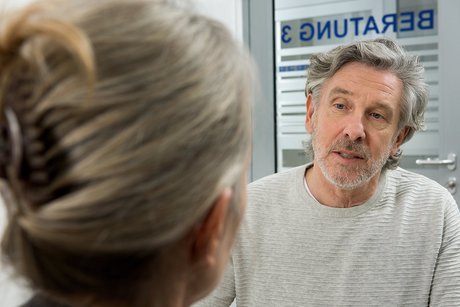 Ein Mann spricht mit einer Frau. Im Hintergrund erkennt man auf einer Glas-Trennscheibe das Wort "Beratung".