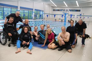 Das Gruppenfoto zeigt die Mitglieder des Mitglieder des 1. Berliner Inklusions-Tauchclubs am Beckenrand im Schwimmbad. 