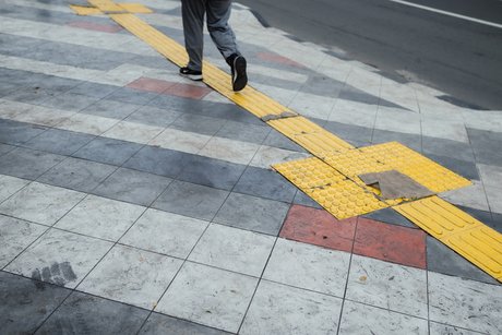 Jemand geht auf einem Gehweg, ein taktiles Blindenleitsystem in gelber Farbe markiert eine Strecke