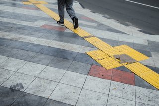 Jemand geht auf einem Gehweg, ein taktiles Blindenleitsystem in gelber Farbe markiert eine Strecke