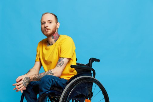Barrierefreiheit Ein junger Mann im gelben T-Shirt, der im Rollstuhl sitzt, vor blauem Hintergrund. Er blickt ernst in die Kamera.