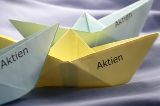 Drei Papierschiffchen, gefaltet aus buntem Papier. Auf den Schiffchen steht jeweils das Wort "Aktien".