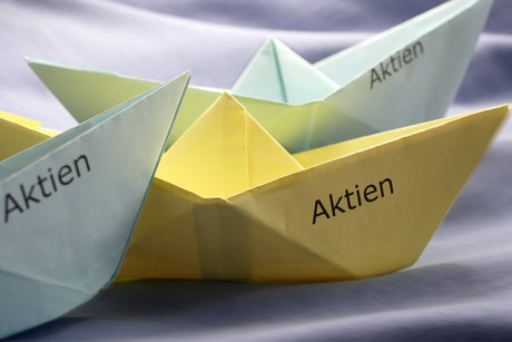 Drei Papierschiffchen, gefaltet aus buntem Papier. Auf den Schiffchen steht jeweils das Wort "Aktien".